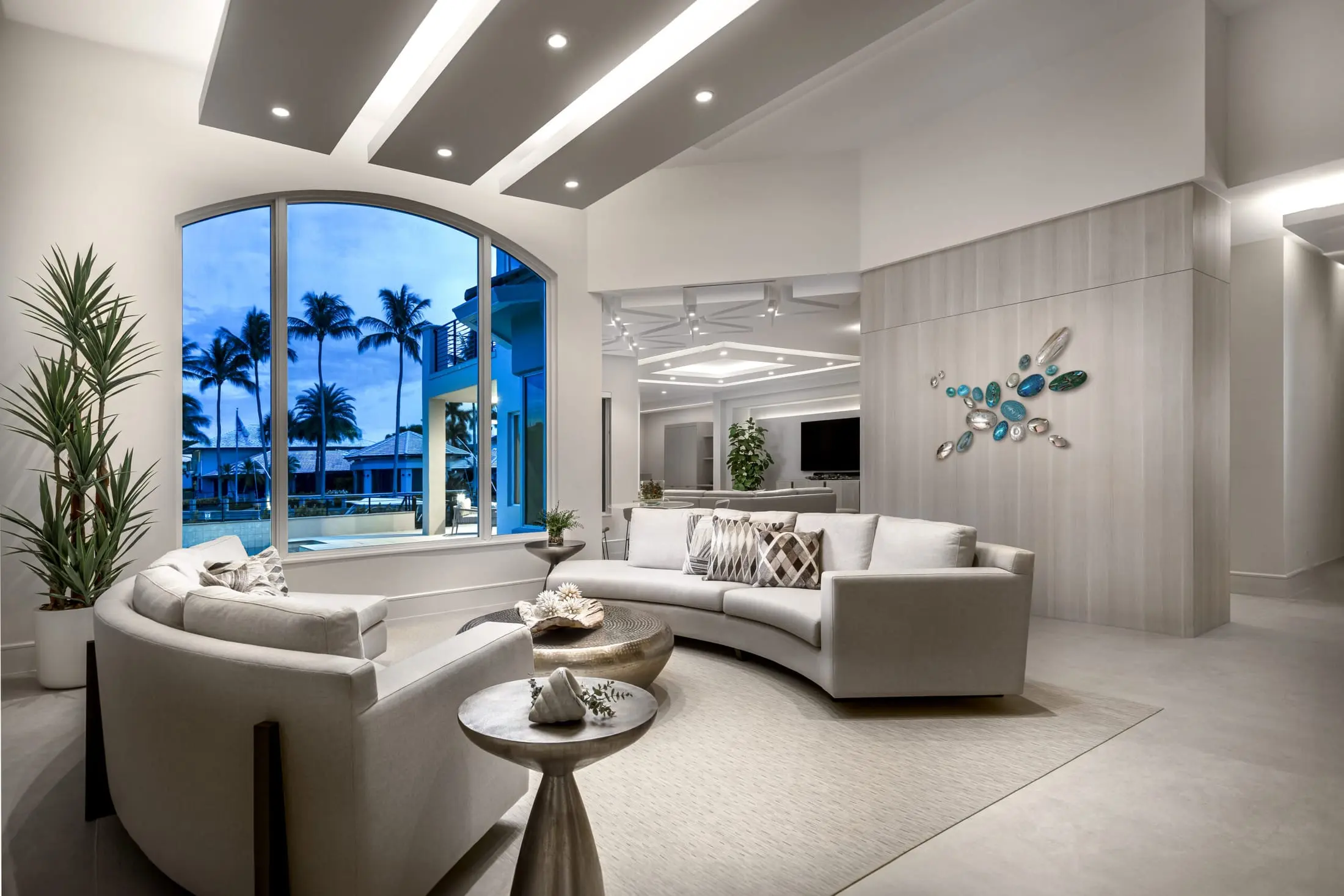 Modern coastal living room with curved sofas, waterfront view, and layered ceiling lighting.