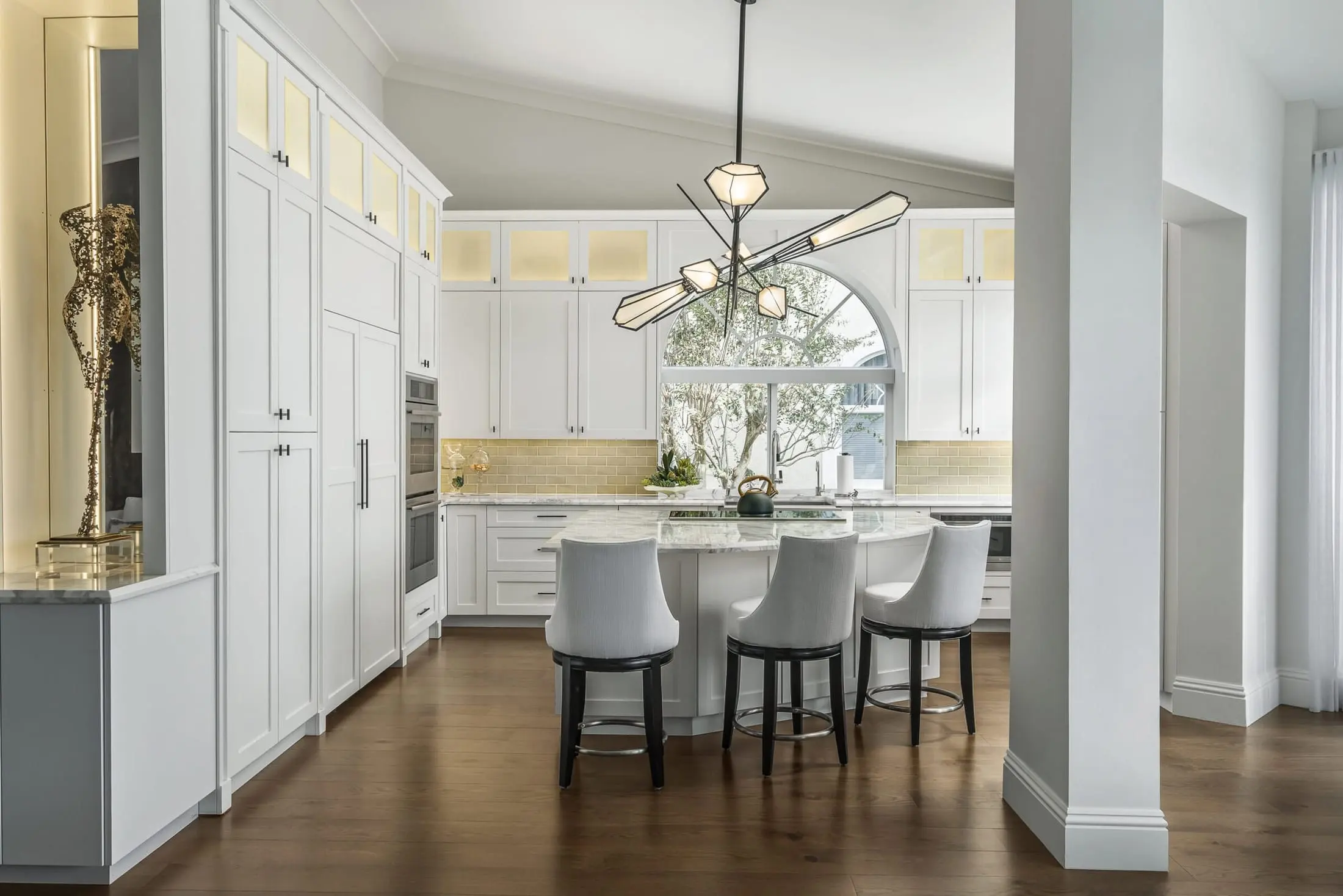 Bright modern kitchen in an Admirals Cove home in Jupiter featuring white cabinetry, marble island with upholstered stools, unique sculptural lighting, and an arched window.