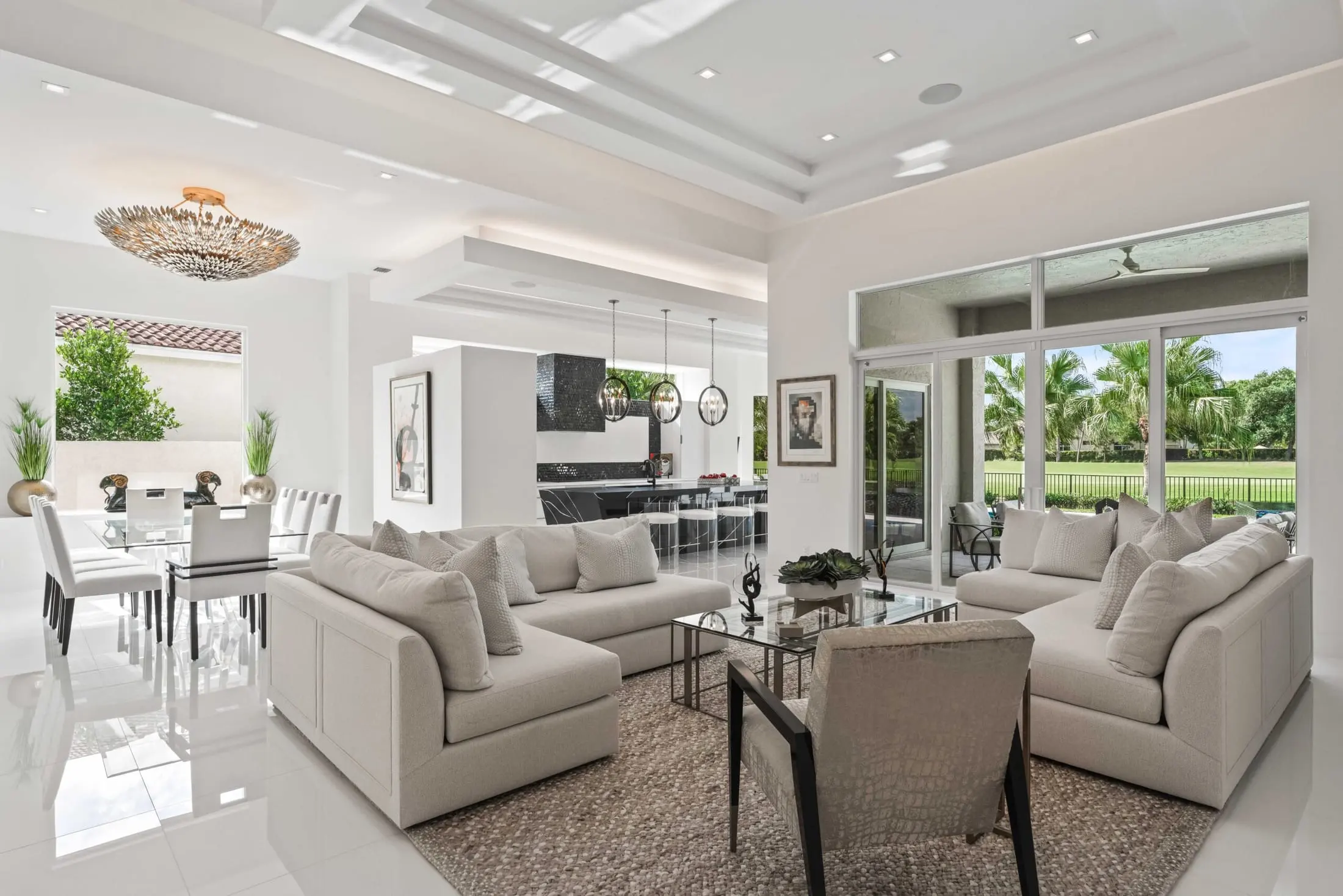 Interior designers Delray Beach Florida - Open living and dining area with neutral sofas, glass dining table, and statement chandelier overlooking the pool.