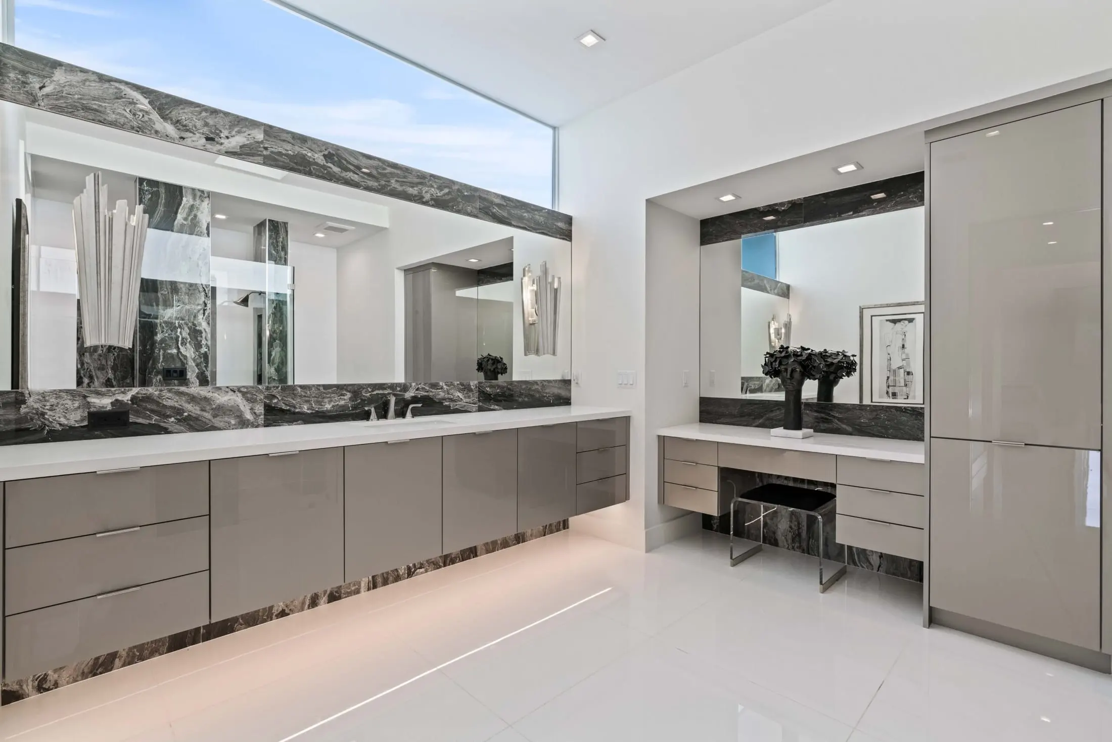 Modern bathroom with sleek taupe cabinetry, marble accents, and a large mirror under natural skylight.