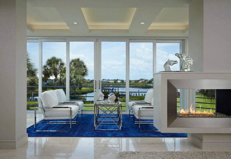 Modern waterfront living room with bold blue rug, sleek fireplace, and white armchairs in Jupiter home.