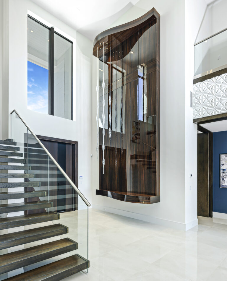 Luxury foyer with floating staircase, glass railing, and sculptural wood wall feature with cascading pendant lighting.