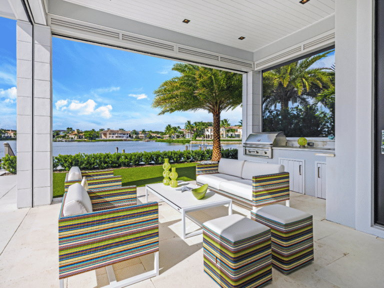 Vibrant outdoor living area in a Palm Beach home featuring striped modern seating, white patio furniture, built-in grill, and waterway views framed by tropical landscaping.