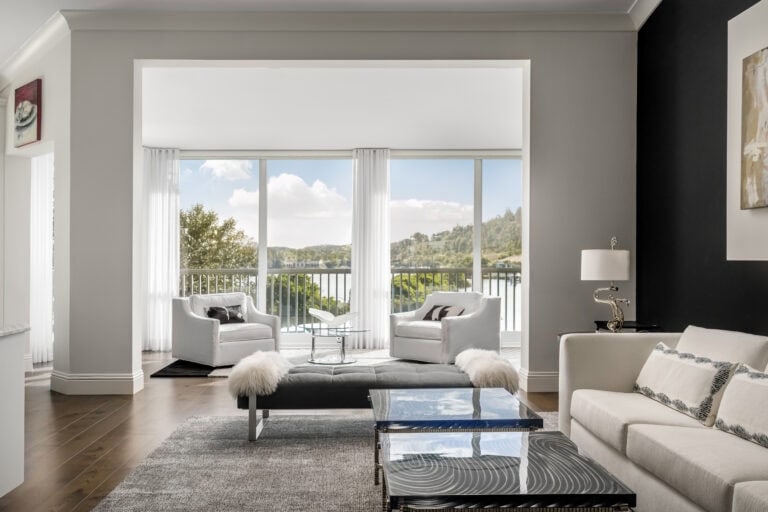 Bright modern living room in an Admiral’s Cove home in Jupiter featuring white lounge chairs, large windows with water views, and contemporary layered furnishings.