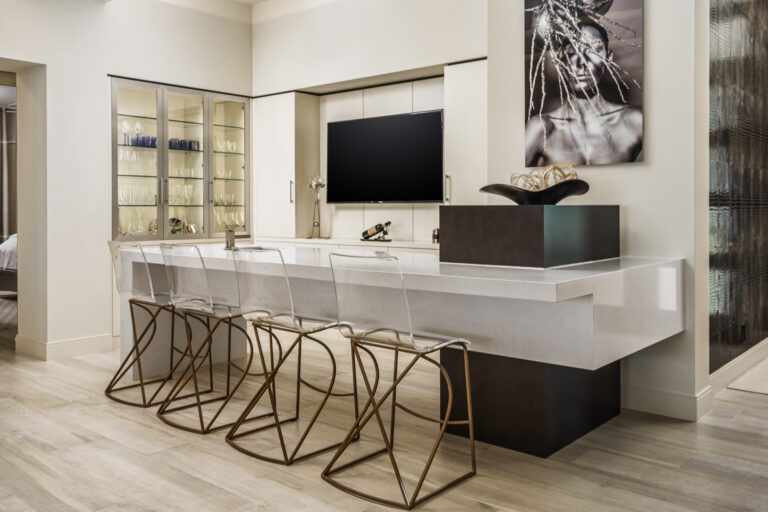 Modern home bar in an Admiral’s Cove residence in Jupiter featuring a sculptural white counter, acrylic barstools with gold bases, built-in glass cabinetry, and contemporary artwork.