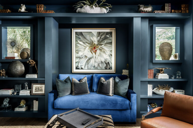Navy built-in shelves framing a bold blue velvet sofa with layered pillows and botanical art in an Admiral’s Cove reading den in Jupiter.
