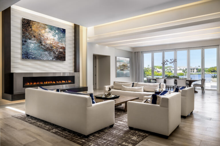 Open-concept living room in an Admiral’s Cove home in Jupiter featuring a sleek linear fireplace, white modern sofas, large windows with waterfront views, and a contemporary dining area.