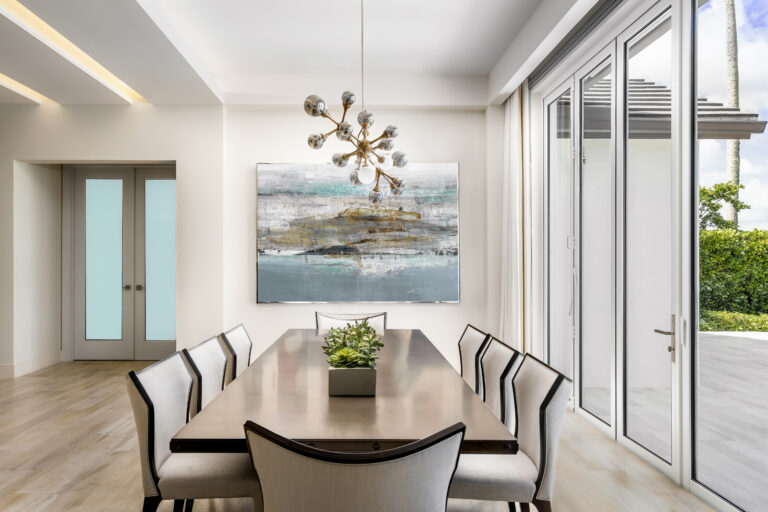 Modern dining room in an Admiral’s Cove home in Jupiter featuring a sleek rectangular table, upholstered chairs, sculptural chandelier, and floor-to-ceiling windows.