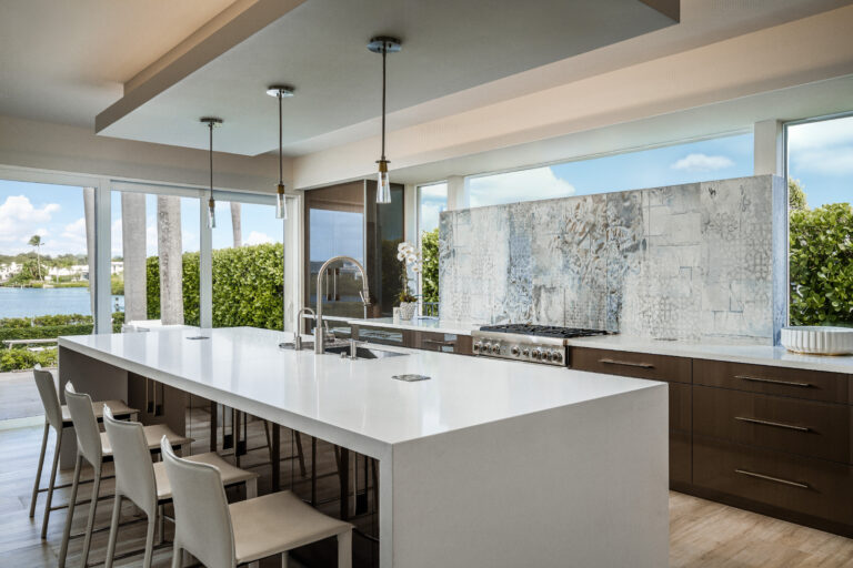 Contemporary kitchen in an Admiral’s Cove home in Jupiter featuring a large white island, sleek cabinetry, textured stone backsplash, and expansive windows with waterfront views.
